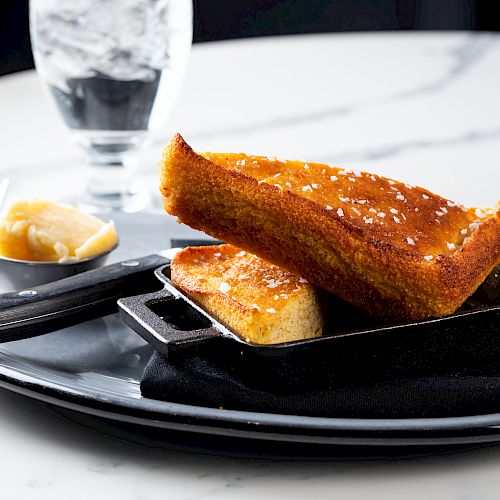 The image shows a black plate with toasted bread topped with coarse salt, a serving of butter, a fork, and a glass of water on a white table.