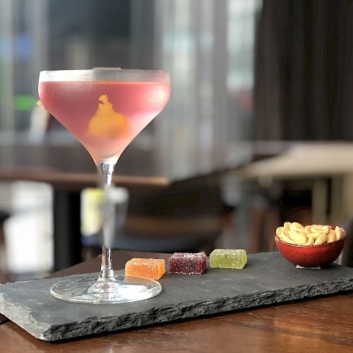 A pink cocktail in a martini glass, garnished with an orange twist, is on a slate with colorful gummies and a small bowl of snacks.