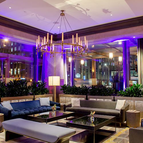 The image shows a stylish lounge area with modern furniture, a chandelier, purple lighting, and city views through large windows, creating a cozy ambiance.