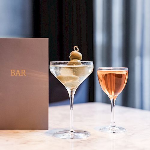 A bar menu is placed next to two cocktails on a marble table, one garnished with olives and the other featuring a rosé colored drink.