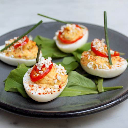 This image shows a black plate with four deviled eggs, each garnished with a slice of red pepper, cheese crumbles, and a chive, resting on green leaves.