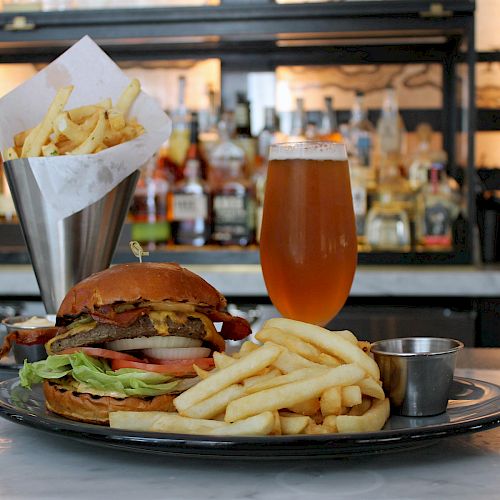 A plate with a burger, fries, and sauce, a cone of fries, and a glass of beer on a counter with a bar in the background.