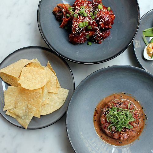 The image shows four dishes: tortilla chips, marinated meat with greens, deviled eggs with garnish, and glazed chicken wings with sesame seeds.