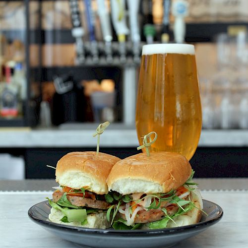 The image shows two sandwiches on a plate, with a glass of beer in the background. The scene is set on a table in a bar or restaurant.