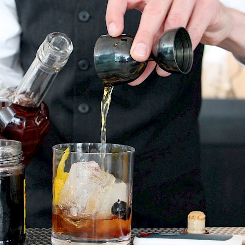 A bartender pouring a drink into a glass with ice and garnishes from a jigger, with a bottle and other bar tools nearby on the counter.