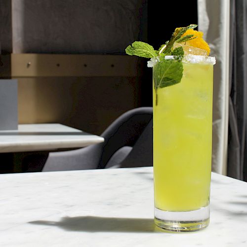 A tall glass filled with a green drink, garnished with mint leaves and an orange slice, sits on a marble table in a well-lit indoor setting.