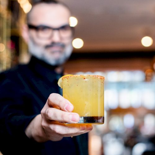 A person holding a cocktail glass with a salted rim and a lemon garnish, set against a blurred bar background.