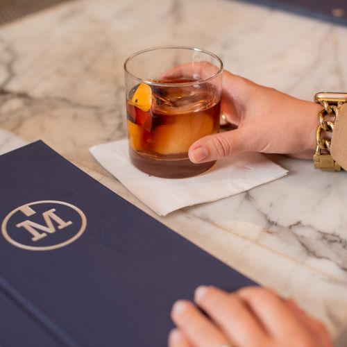 A person holds a drink with an orange garnish on a marble table next to a blue menu with a circular "M" logo.