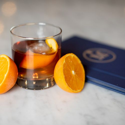 A glass with a cocktail, garnished with an orange peel, sits on a marble surface, surrounded by orange halves and a blue menu.