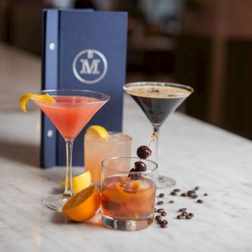 A variety of cocktails on a marble surface, with garnishes and a blue menu in the background.