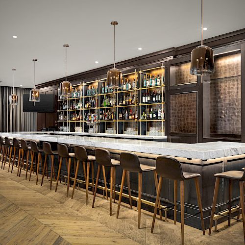 The image shows a modern bar with a long marble countertop, wooden barstools, shelves stocked with various bottles, and pendant lighting.