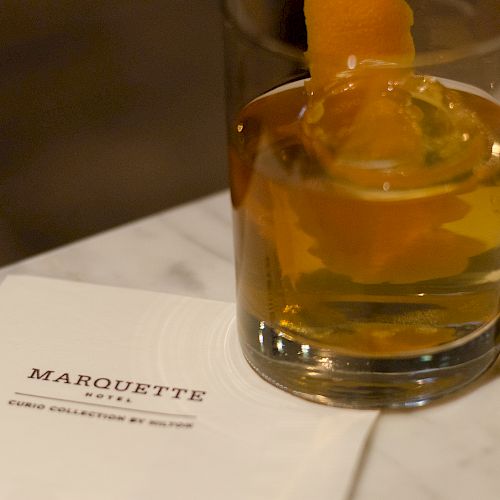 A glass with a beverage and an orange peel alongside a "Marquette Hotel" napkin on a marble surface.