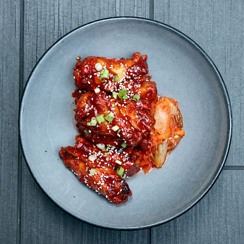 A plate of spicy glazed chicken wings garnished with sesame seeds and green onions, on a gray slate surface.