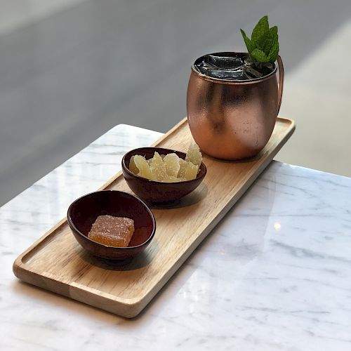 A decorative wooden tray with a copper mug with a mint garnish, a bowl of sugar cubes, and a bowl with a brown sugar cube on a marble surface.