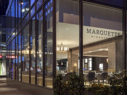 A modern building with large glass windows displaying "Marquette Minneapolis" and a sign for "Marquette Hotel" under a skybridge at night.