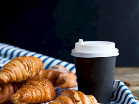 The image shows a few croissants on a plate, a takeaway coffee cup with a white lid, and coffee beans scattered on a wooden table with a striped cloth.