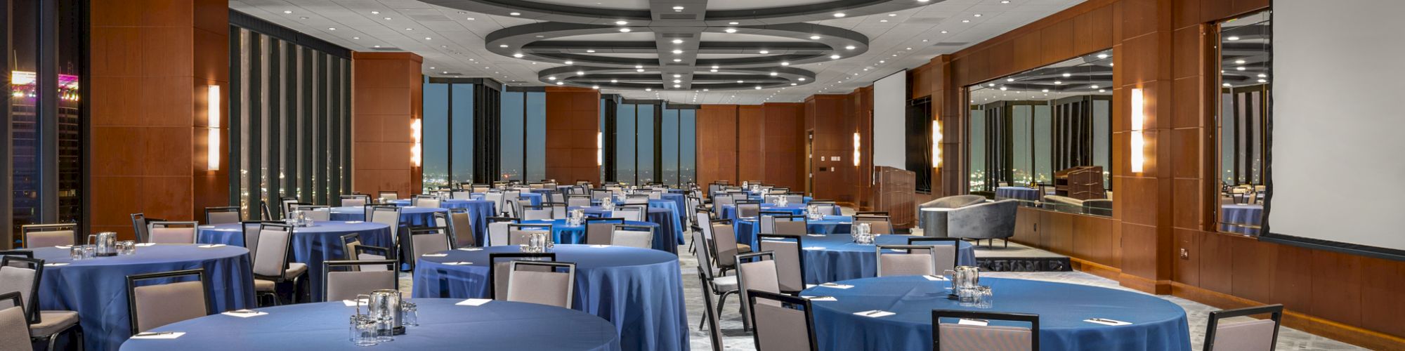 The image shows a spacious conference or banquet room with circular tables covered in blue tablecloths and surrounded by chairs, under modern lighting.