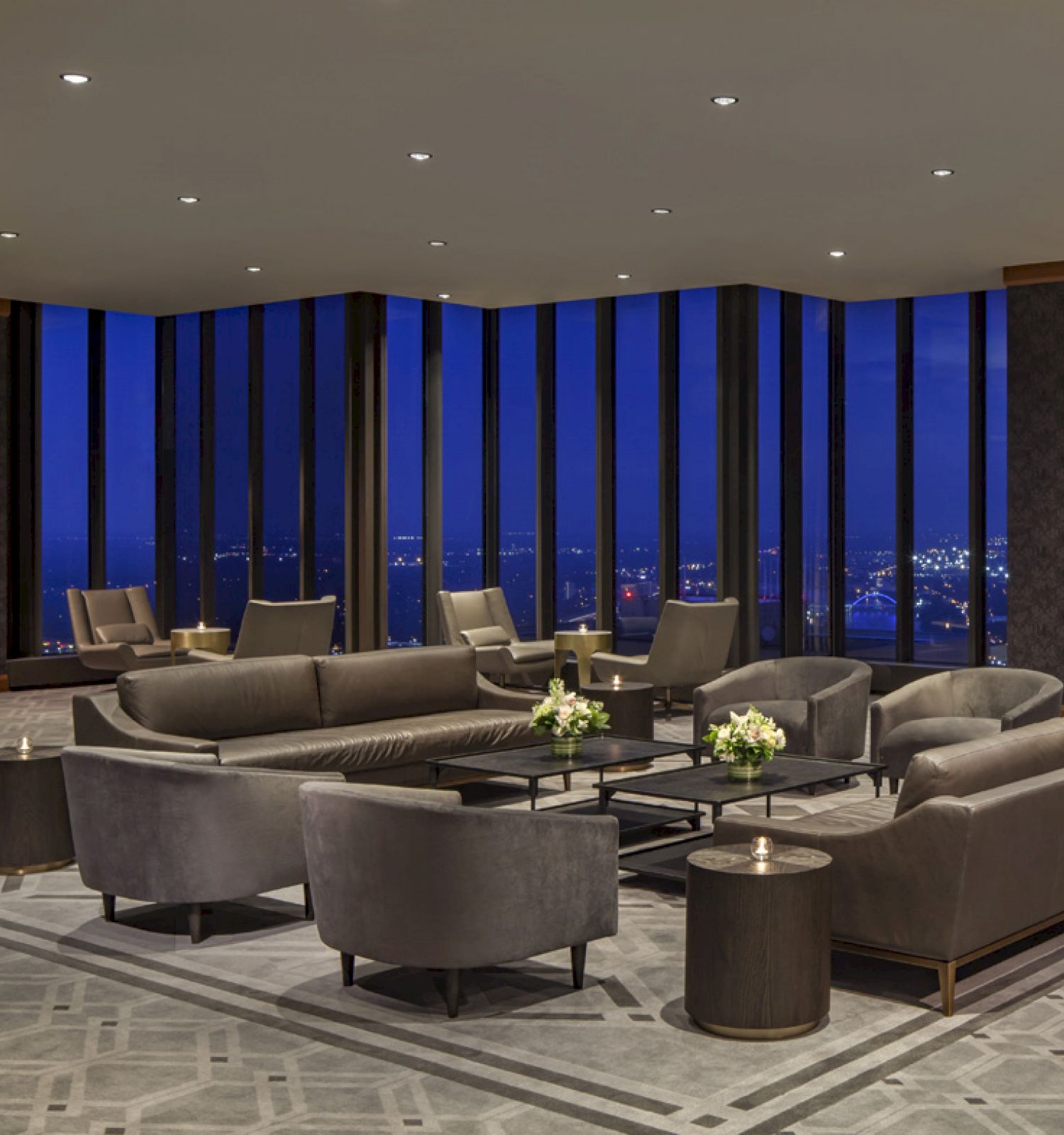 The image showcases an elegant, modern lounge with large windows, gray sofas, armchairs, and a nighttime city view, providing a luxurious atmosphere.