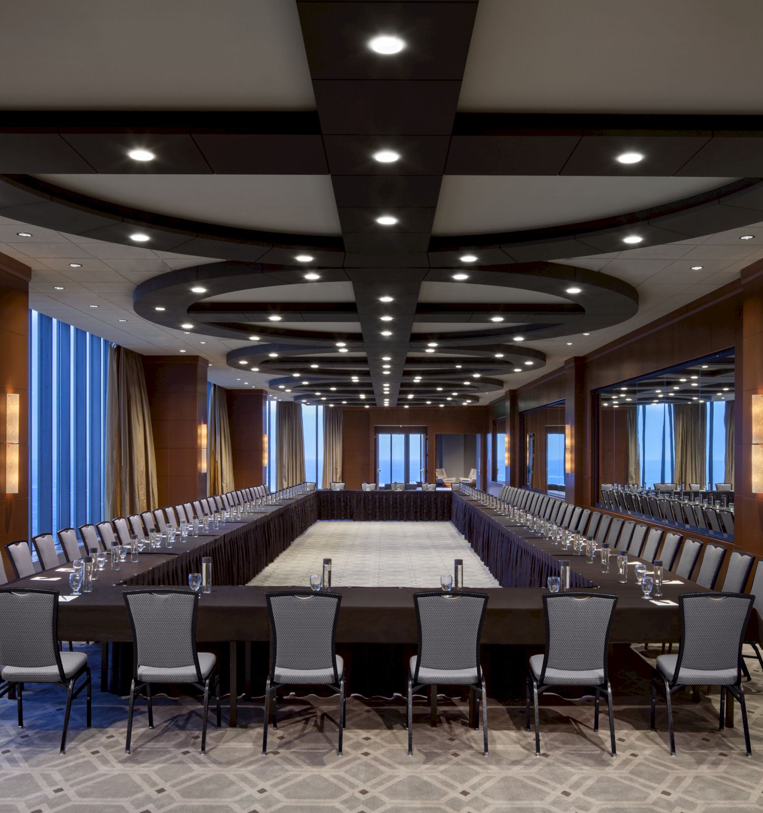 A large, modern conference room with an empty U-shaped table setup, surrounded by numerous chairs and decorated with wood paneling and large mirrors.