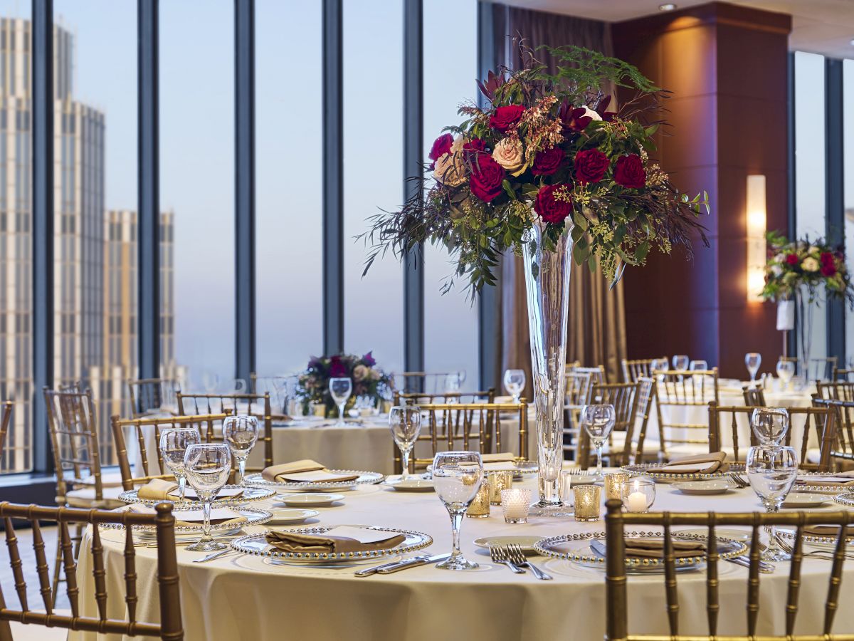 A beautifully set banquet room with round tables, elegant floral centerpieces, gold chairs, and large windows offering a city skyline view in the background.