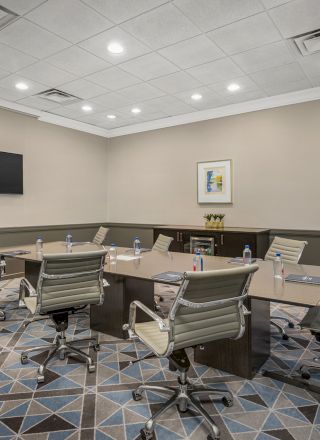 The image shows a modern conference room with a rectangular table, swivel chairs, a wall-mounted TV, a painting, and water bottles on the table.