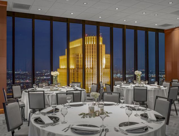 A formal dining room with round tables set for an event is overlooking a night skyline with a prominent illuminated building in the background.