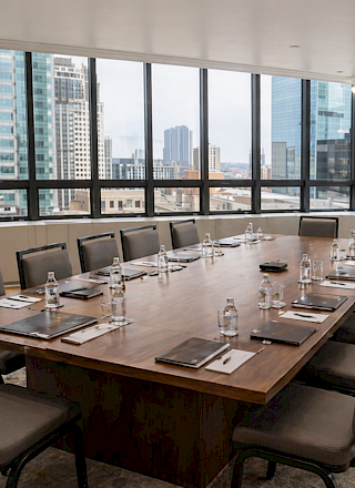 A bright city skyline meeting room with a long wooden table set for a meeting, water bottles, glasses, and notebooks. 140 characters.