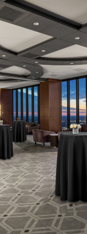 A spacious and elegant event room with large windows offering a sunset view, decorated with round tables covered in black cloth, and modern ceiling lights.