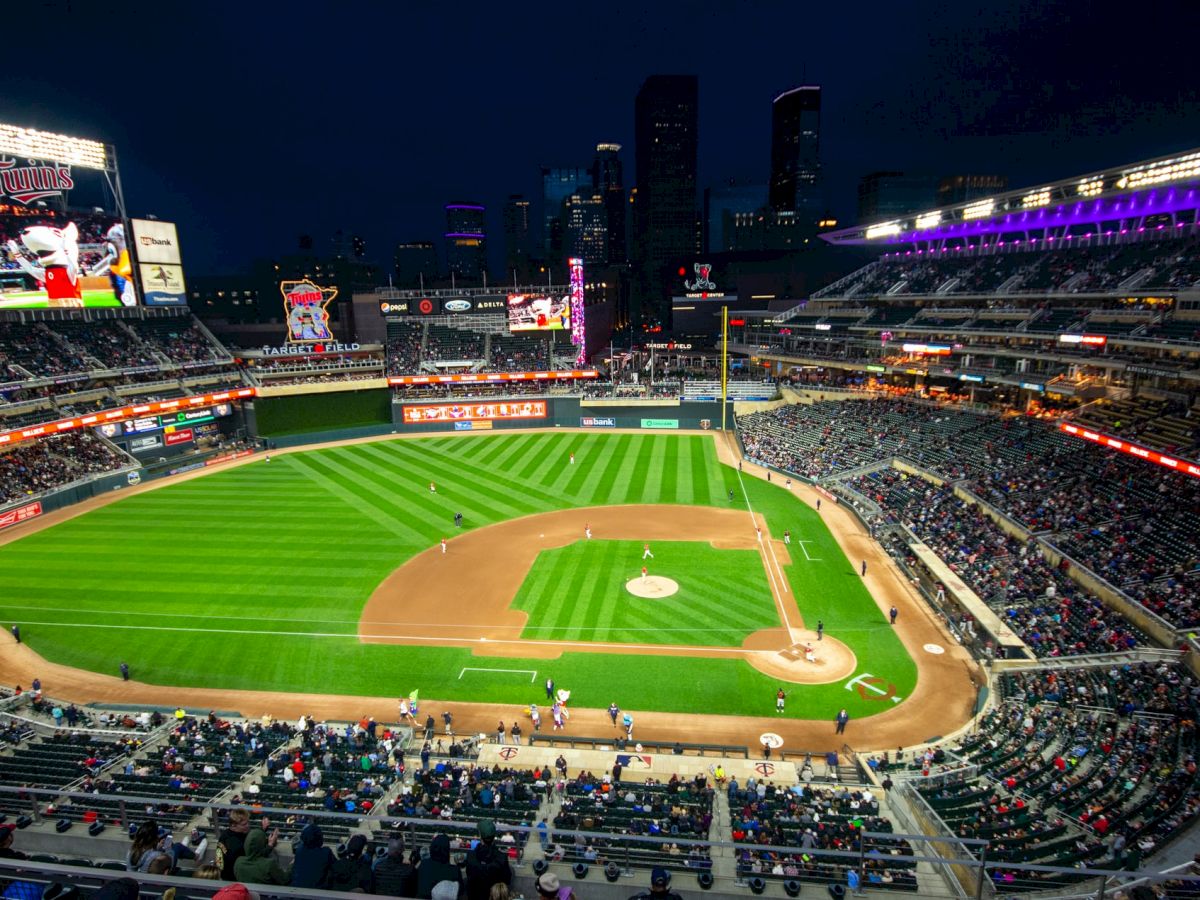 A nighttime baseball game is underway in a large stadium with fans in the stands and a city skyline visible in the background.