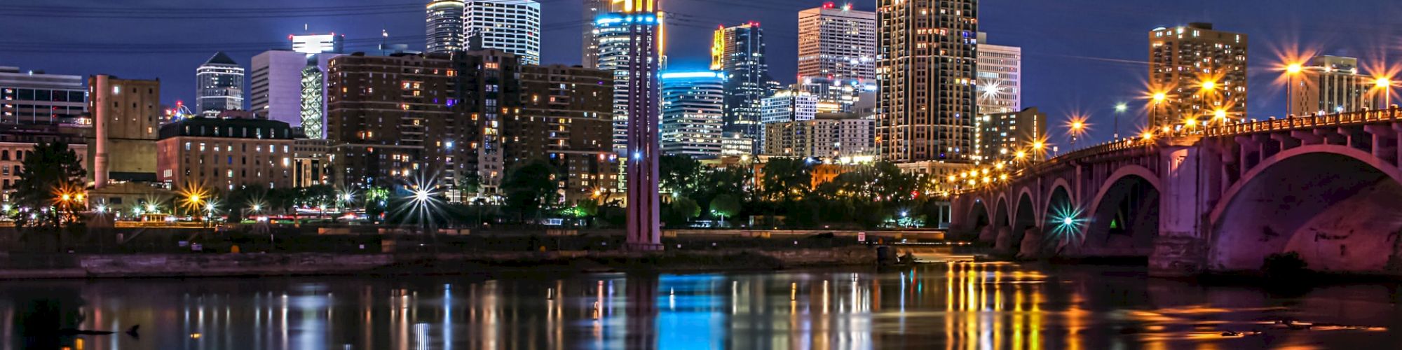 A city skyline at night with illuminated buildings reflecting on the water, and a brightly lit bridge on the right side of the image.