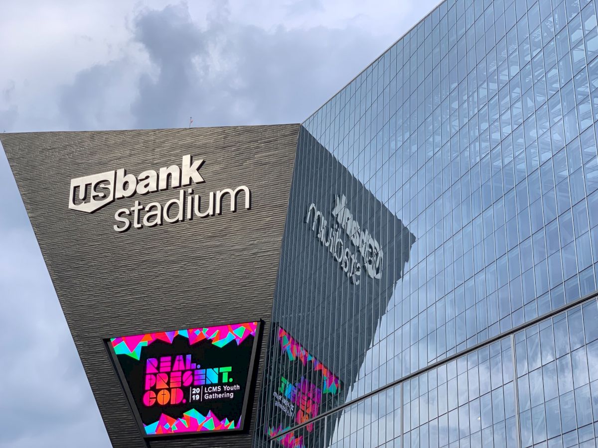 Image of the exterior of US Bank Stadium. The building has reflective glass surfaces and electronic signage displaying colorful text.