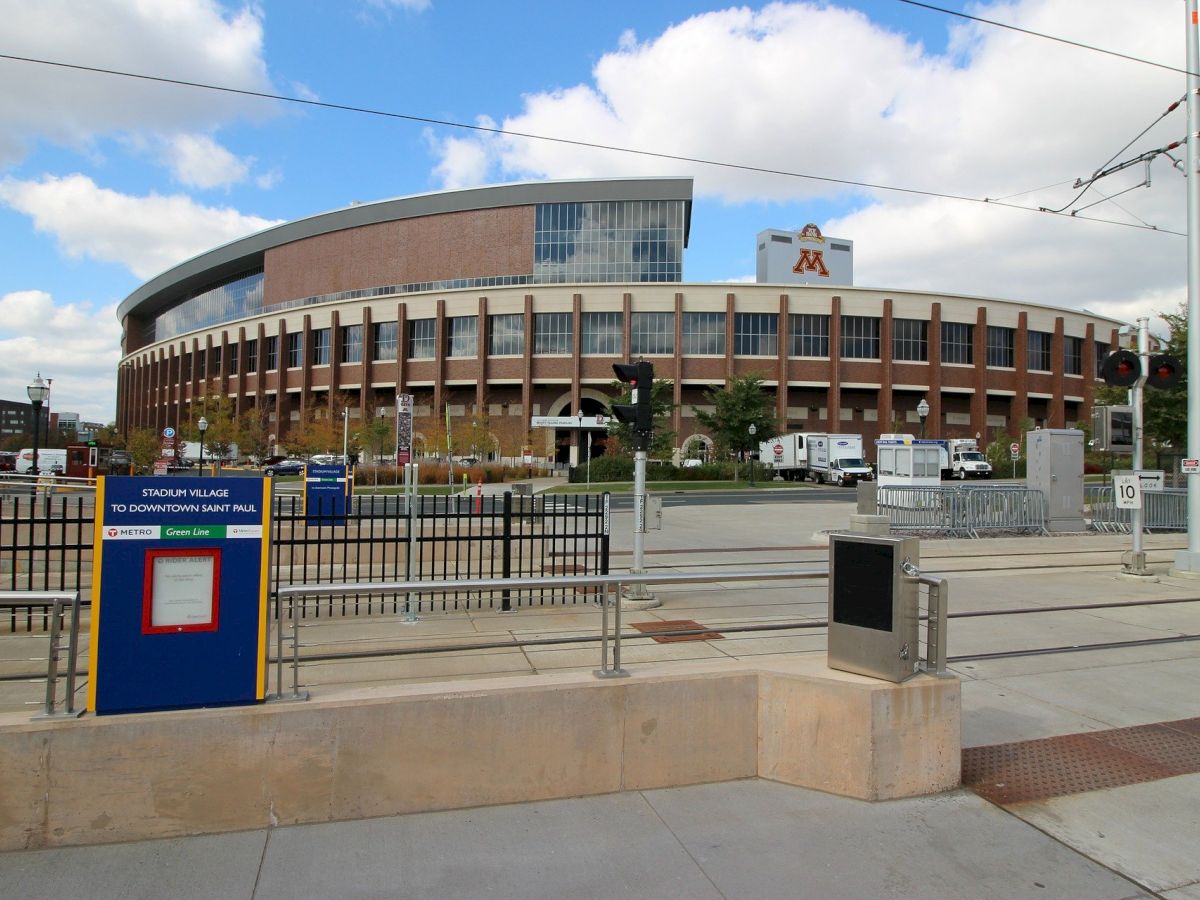 An exterior view of a large, oval-shaped building with a sign that reads "M," likely a sports or event arena, with a railway or tram track in the foreground.