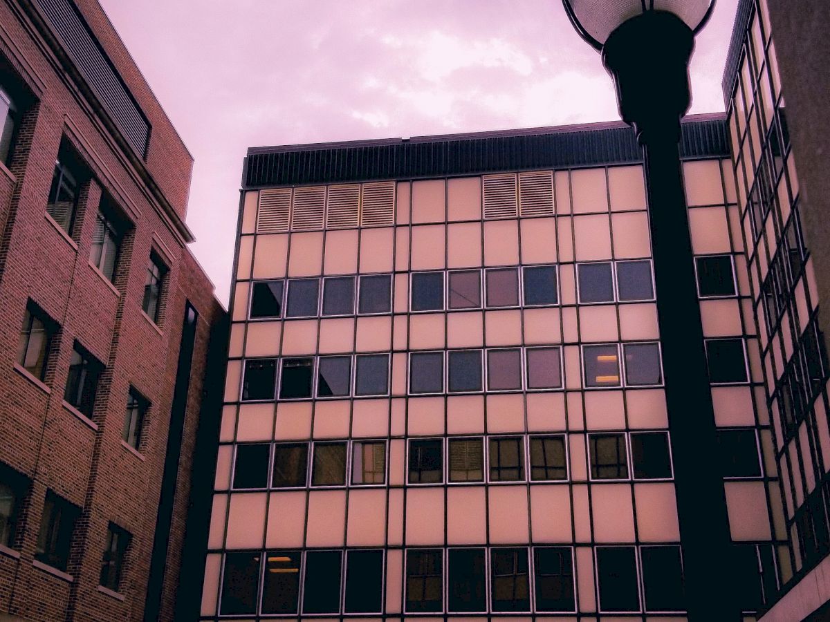 The image shows a multi-story building with a grid-like window design, framed by two other buildings and a streetlamp in the foreground, against a purple sky.