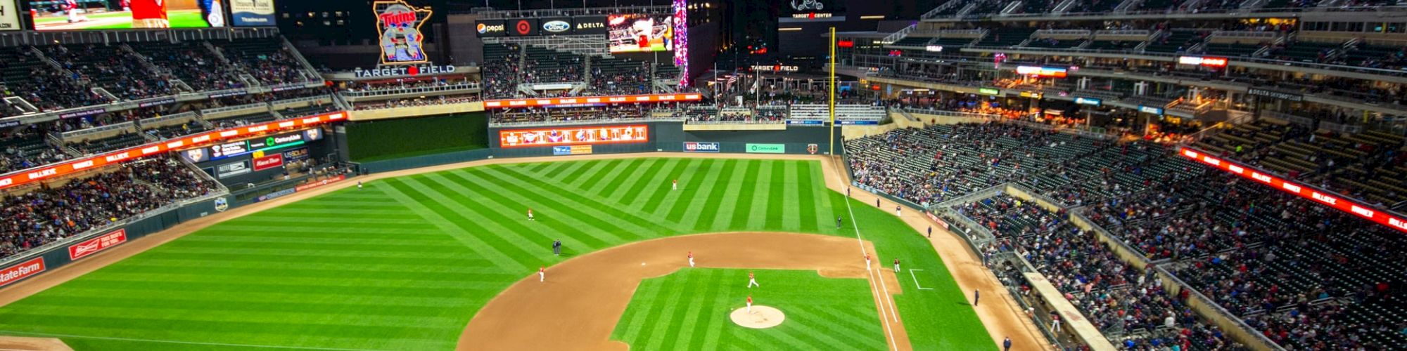 A nighttime baseball game is taking place in a well-lit stadium with spectators in the stands and the city skyline in the background.