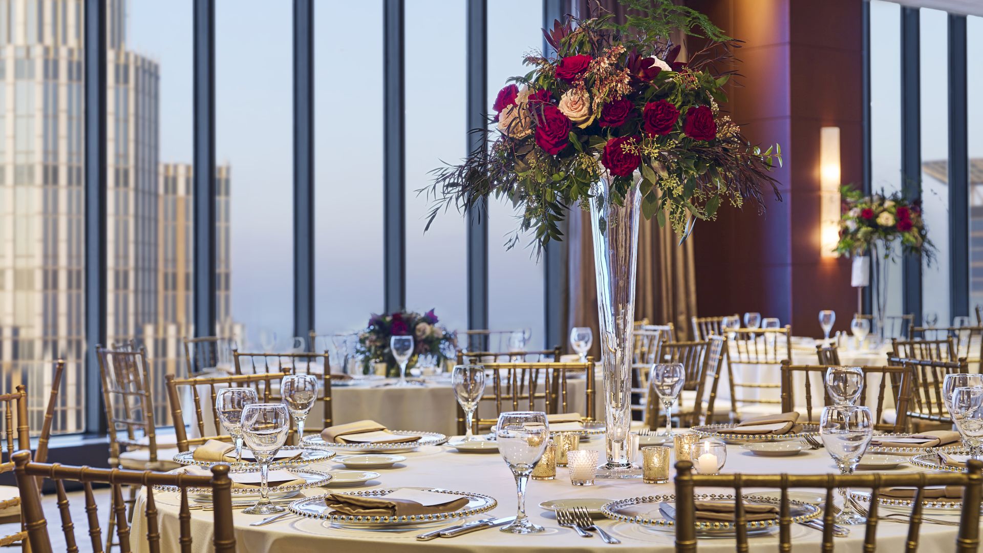 A beautifully decorated banquet hall with large windows, featuring elegant table settings and floral centerpieces, is seen in the image, which ends here.