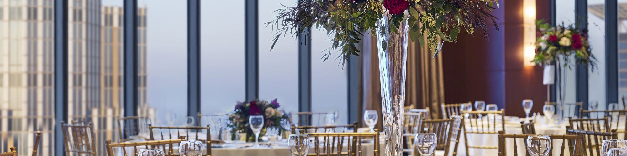 A beautifully decorated banquet hall with large windows, featuring elegant table settings and floral centerpieces, is seen in the image, which ends here.