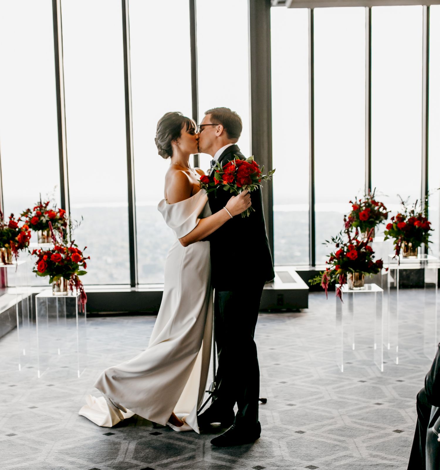 A couple is kissing at their wedding ceremony with floral arrangements, held in a modern room with large windows, observed by a few seated guests.