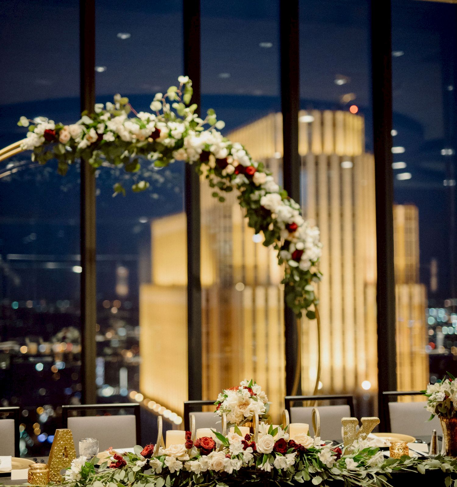 A wedding reception setup with a flower-covered arch and "Mr" & "Mrs" signs on the table, overlooking a cityscape through large windows at night.