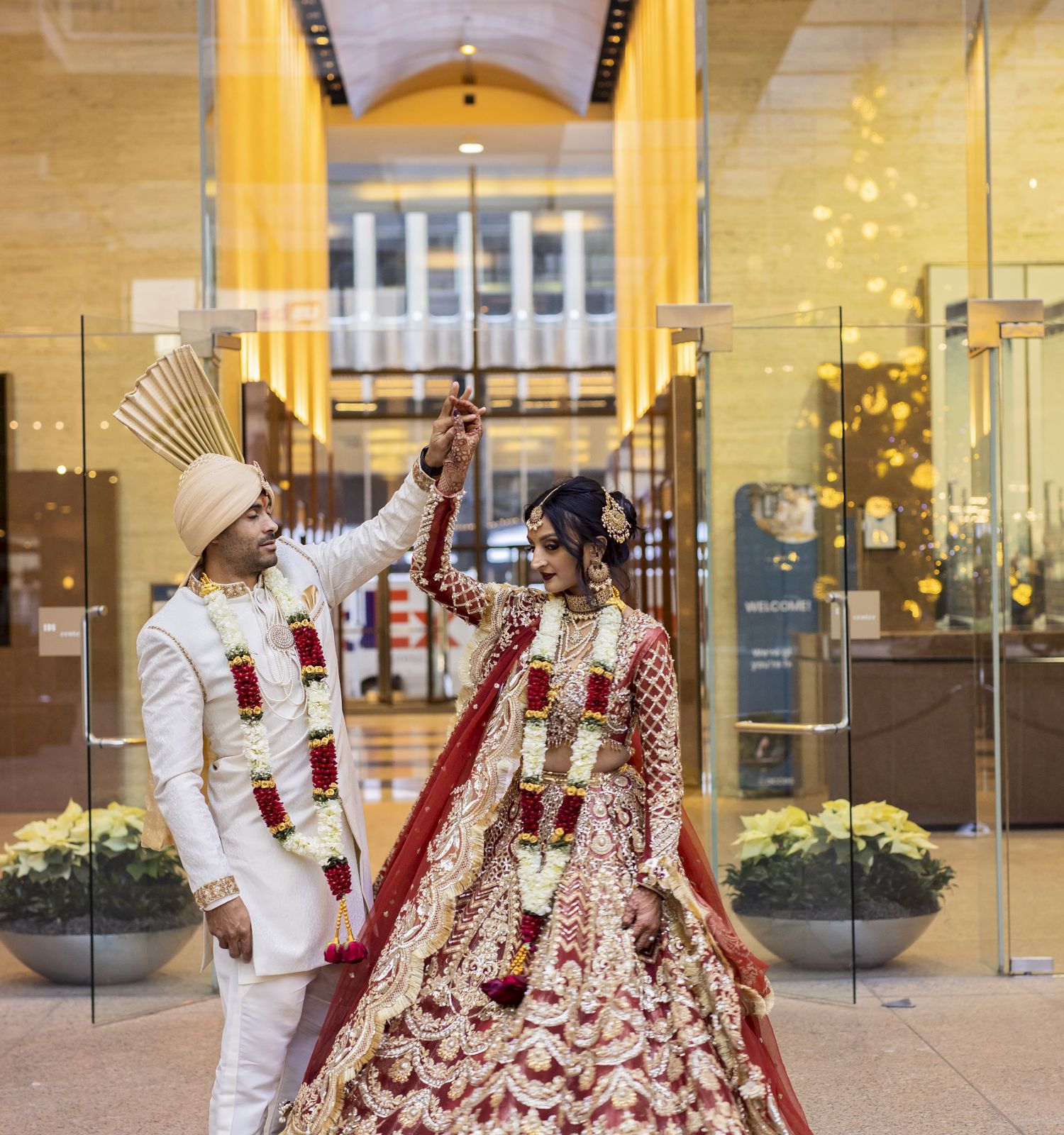 A couple in traditional attire pose inside a modern building with gold accents and large glass doors, the man twirling the woman's hand.