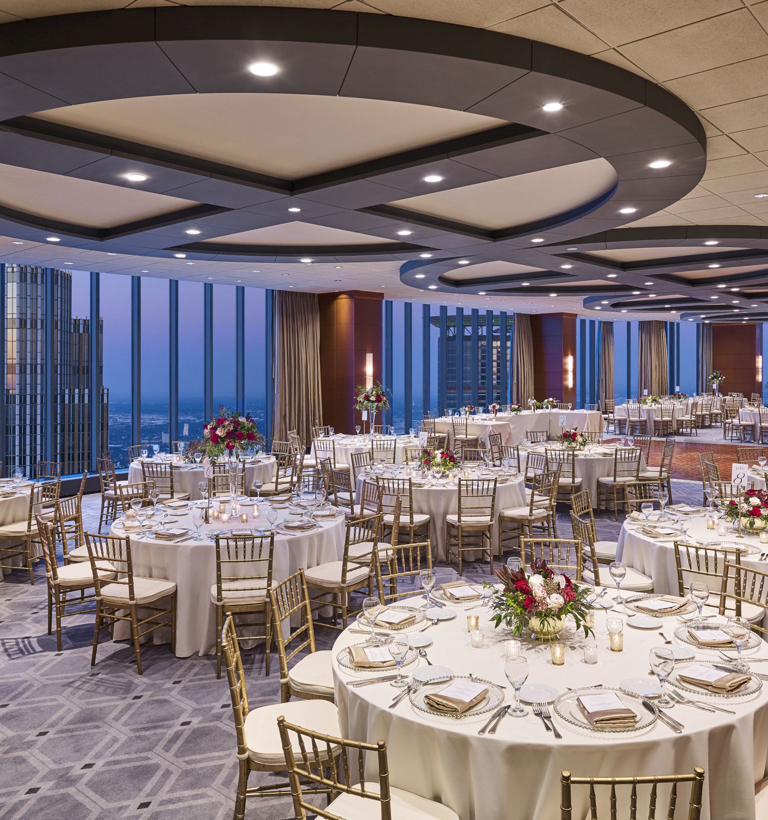 A beautifully decorated banquet hall with round tables, elegant chairs, floral centerpieces, and panoramic city views through tall windows, set for an event.