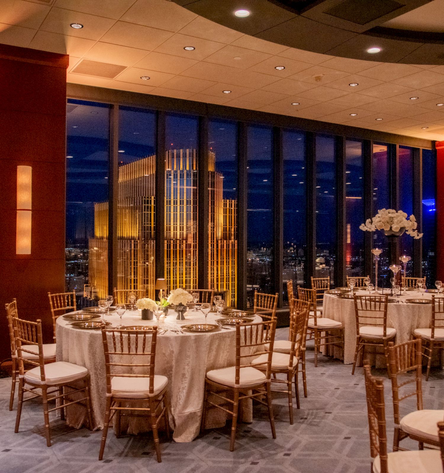 The image shows a beautifully set dining area with round tables, elegant chairs, and a cityscape view through large windows at night.