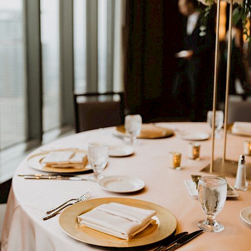 A round table is elegantly set for a formal event with plates, glasses, utensils, napkins, and a floral centerpiece, next to large windows.