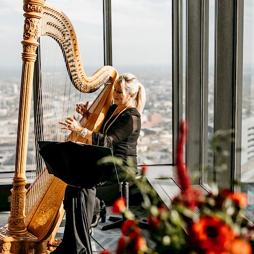 A woman is playing a harp in a room with large windows offering a city view. Vibrant red flowers are in the foreground, slightly out of focus.