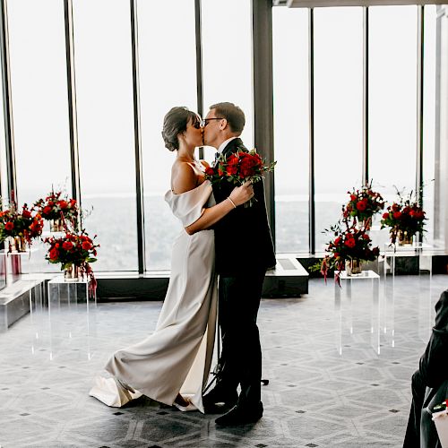 A couple is sharing a kiss in a wedding ceremony with floral decorations and large windows in the background, while guests are seated watching them.