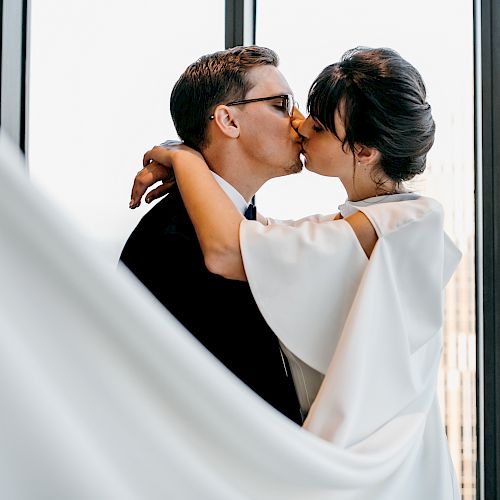 A couple in formal attire kissing in front of cityscape window panels.