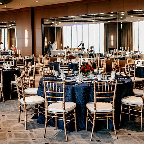 A banquet hall is set up with round tables covered in dark tablecloths, gold chairs, elegant centerpieces, and large windows in the background.