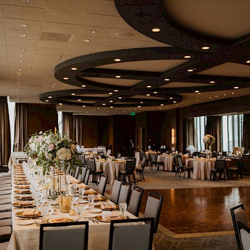 A beautifully decorated banquet hall with a long dining table featuring floral centerpieces, surrounded by chairs and other round tables.