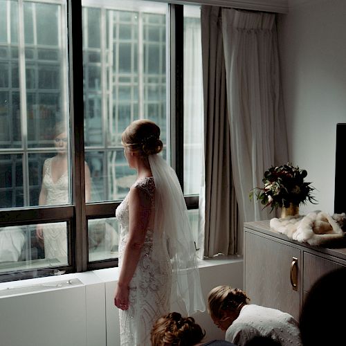 A bride stands facing a window, her reflection visible. Two people are kneeling behind her in a room with a TV and a bouquet on a shelf.