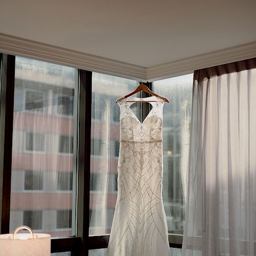 A white wedding dress hangs on a wooden hanger in front of a large window with city buildings in the background.