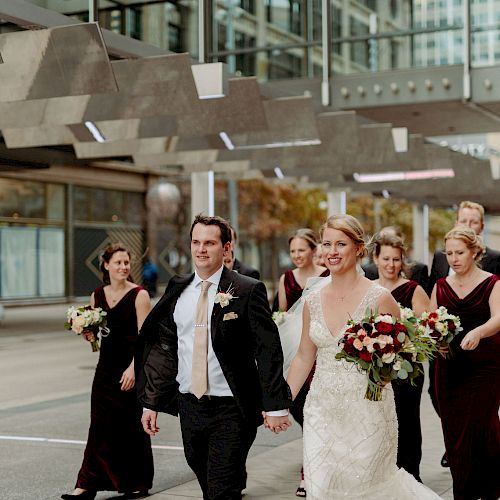 A bride and groom walk hand-in-hand with their bridal party wearing elegant dresses and suits, holding bouquets in an urban setting.